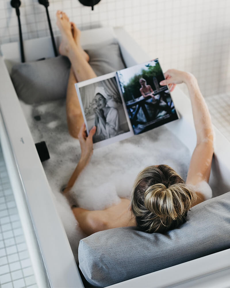 Frau badet mit Rückenkissen (Medium) und Fusskissen in luxuriöser freistehender Badewanne. Rücken, Kopf und Füsse ruhen entspannt auf Badesofas (Farbe Grau). $ Innenform eckig | Size Medium Set | Cloud / Hellgrau