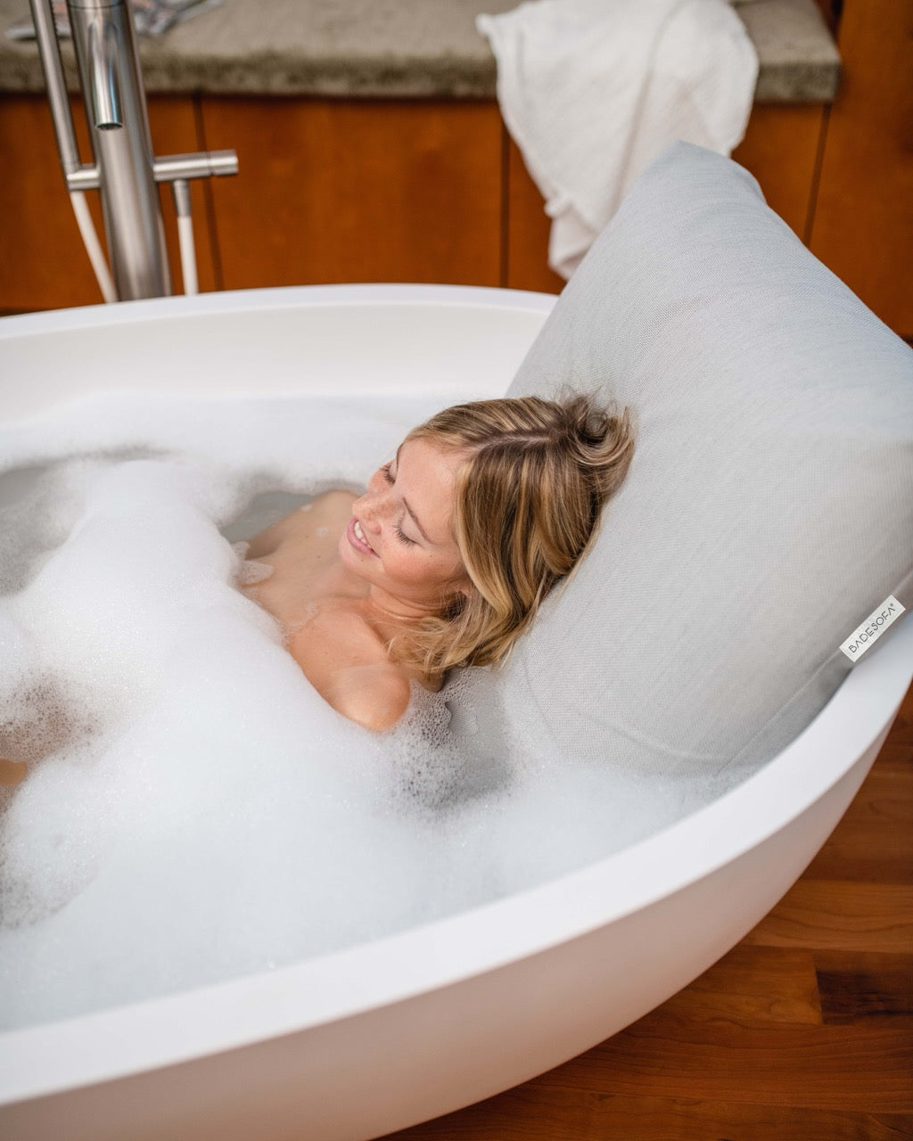Hellgraues Badesofa Rückenkissen (Size Large) in einer freistehenden Badewanne, mit lächelnder Person, die sich entspannt anlehnt.