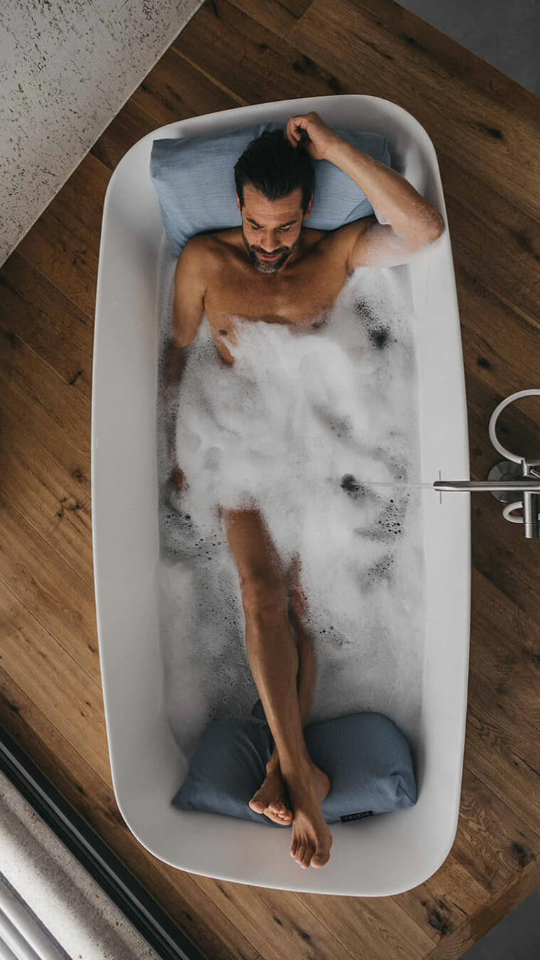 Mann badet mit Rückenkissen (Large, Sky oder Hellblau) und Fusskissen in luxuriöser freistehender Badewanne. Rücken, Kopf und Füsse ruhen entspannt auf Badesofas (Farbe Sky oder Hellblau). $ Freistehende Badewanne | Size Large Set | Sky