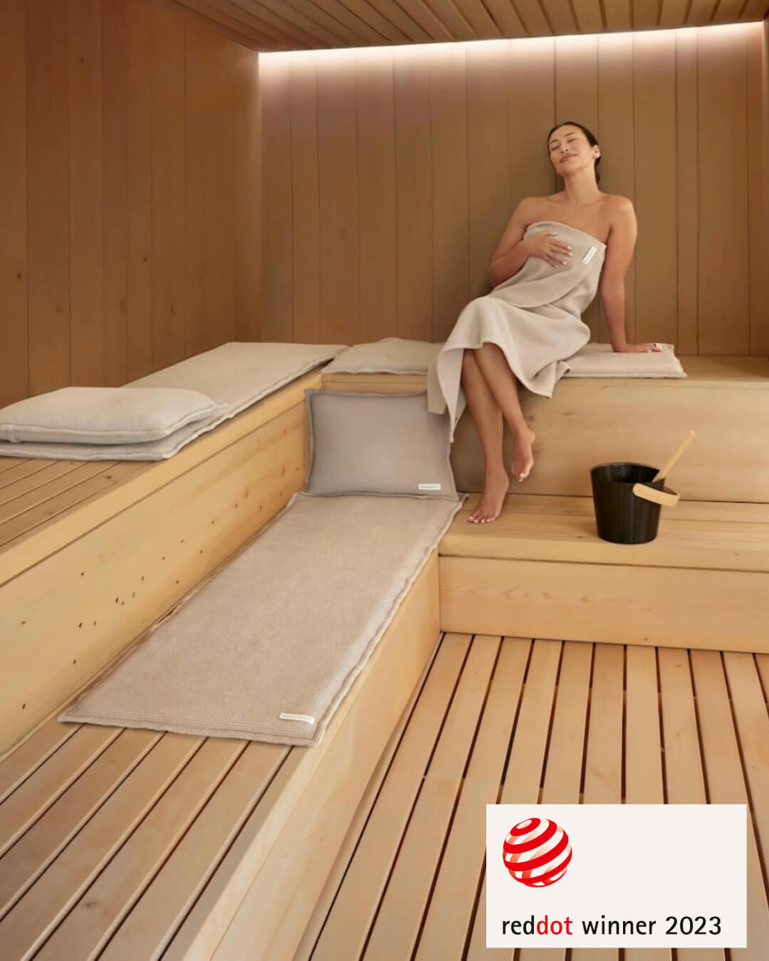 Frau sitzt auf einer Saunaauflage (Größe Short) in einer Sauna, in der zusätzlich zwei Saunasofa Sets in der Größe long liegen. Saunasofa ist Red Dot Award Winner 2023.