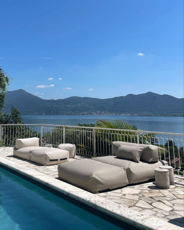 Eine Frau liegt entspannt auf einem Poolsofa double in Beige, das im Pool schwimmt. Am Rand des Pools, auf der Terrasse steht ein weiteres Poolsofa. 