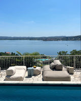 An einem Pool stehen zwei Poolsofas (ein Double in Biege, ein Single in Perlmutt) gegenüber als Outdoorlounge und ein Pouf in Small und wird als Tisch genutzt. 