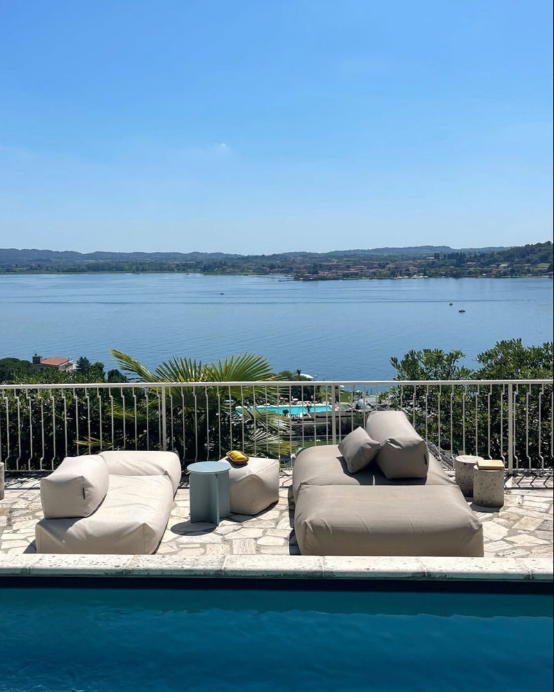 An einem Pool stehen zwei Poolsofas (ein Double in Biege, ein Single in Perlmutt) gegenüber als Outdoorlounge und ein Pouf in Small und wird als Tisch genutzt. 