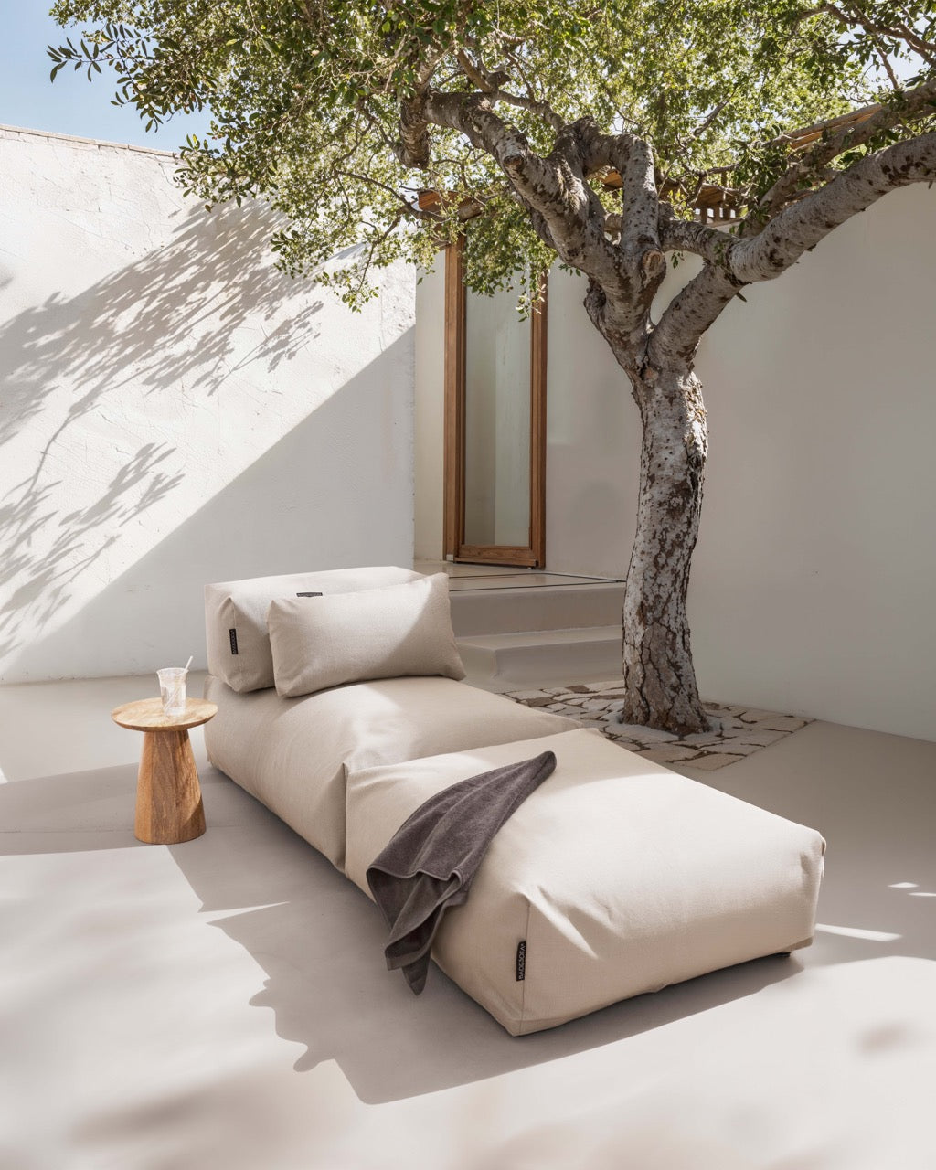 Unter einem Baum steht ein Poolsofa Singel in Beige, daneben ein kleiner Holztisch. Das Rückenkissen und Relaxkissen sind auf dem Poolsofa drapiert.