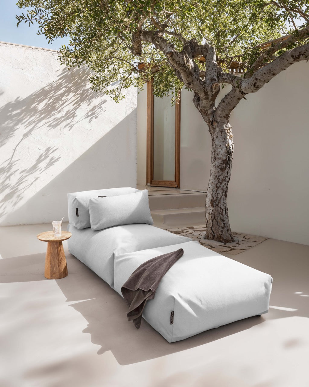Unter einem Baum steht ein Poolsofa Singel in Grau, daneben ein kleiner Holztisch. Das Rückenkissen und Relaxkissen sind auf dem Poolsofa drapiert.